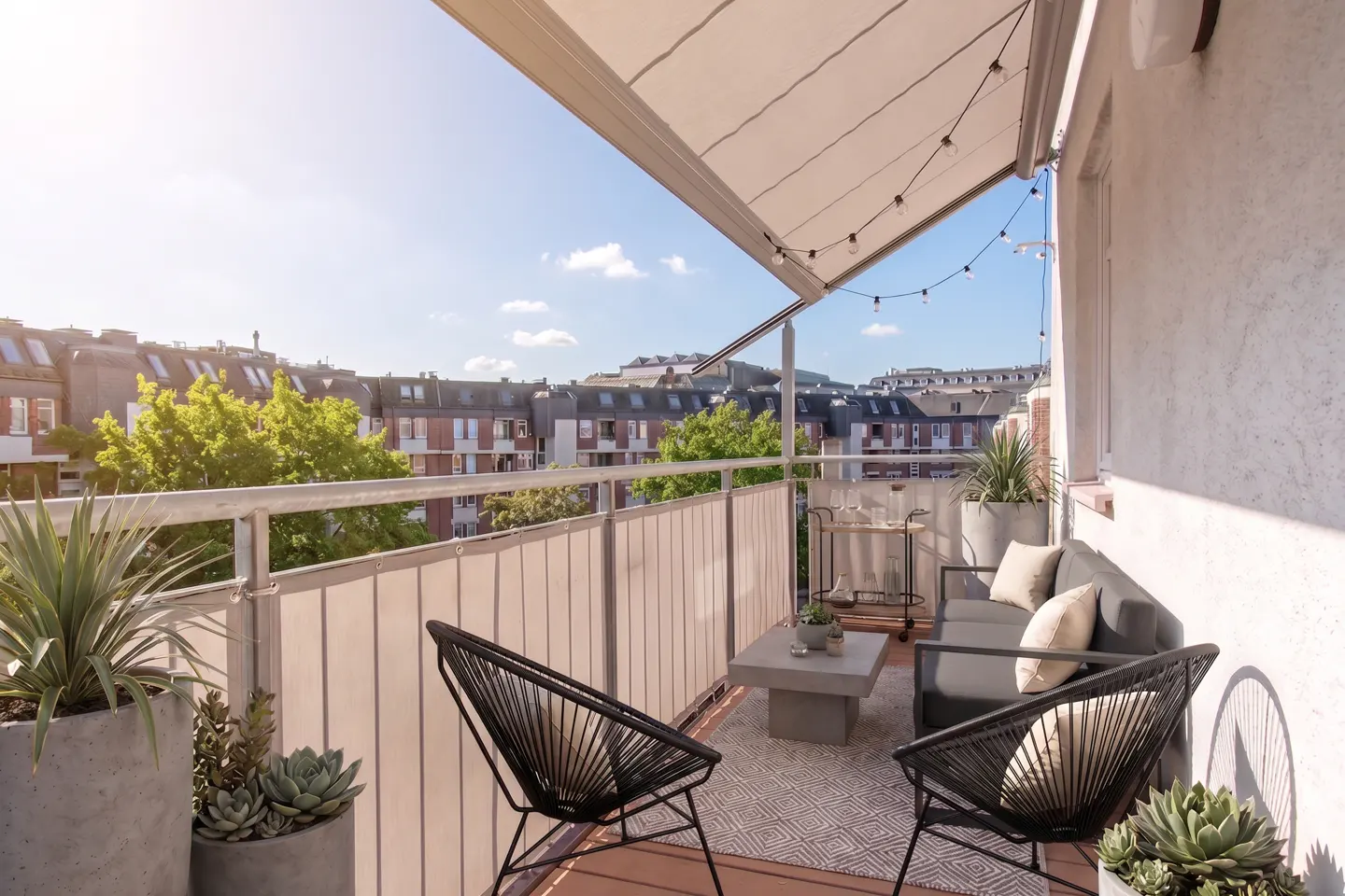 A cozy balcony with black chairs, a gray sofa, and a concrete table. String lights hang under the awning, with a city view in the background.