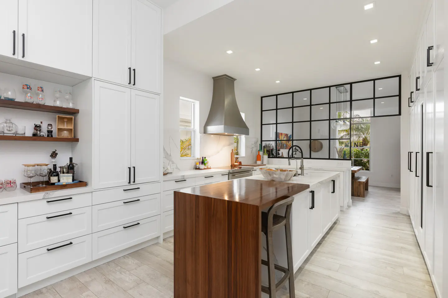 Bright, modern kitchen with white cabinets, black hardware, and wood accents. Island with seating, stainless steel hood, and black-framed windows.