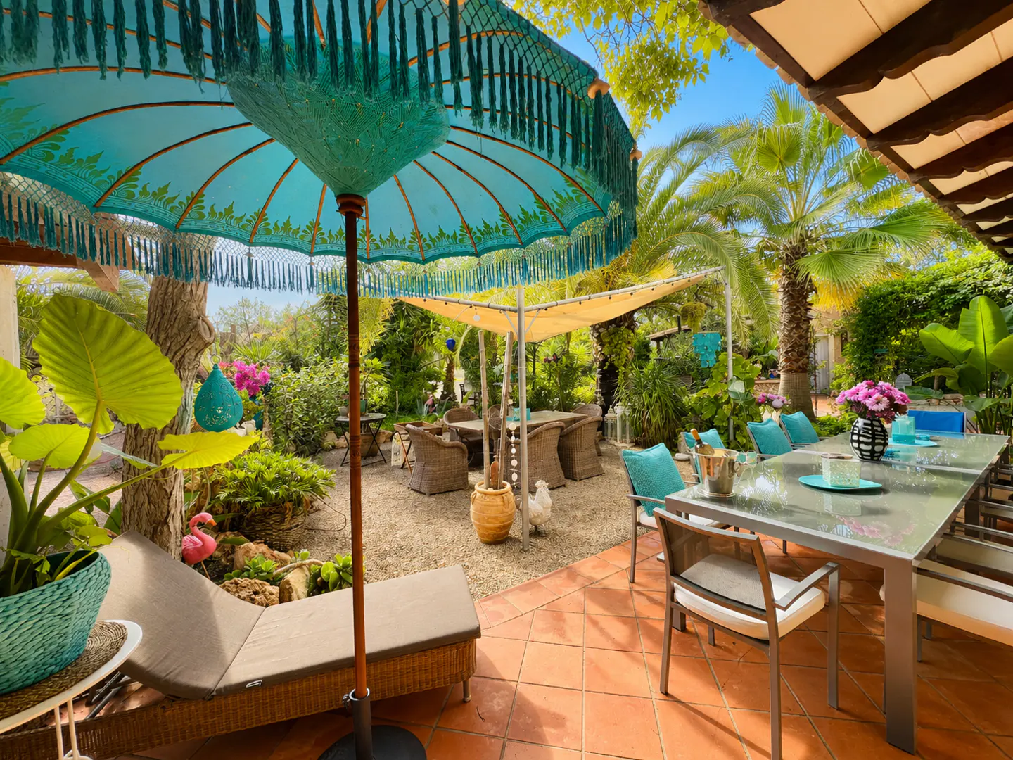 Outdoor patio with a turquoise umbrella, lounge chair, and dining table. Lush greenery and palm trees surround the space.