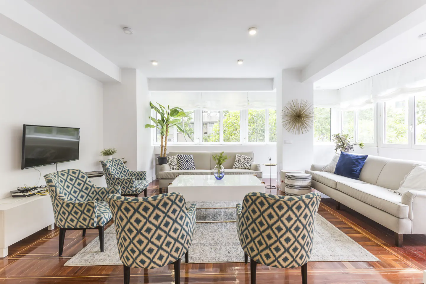 Bright living room with hardwood floors, white walls, and large windows. Four patterned armchairs face two sofas and a white coffee table. A TV is mounted on the wall.