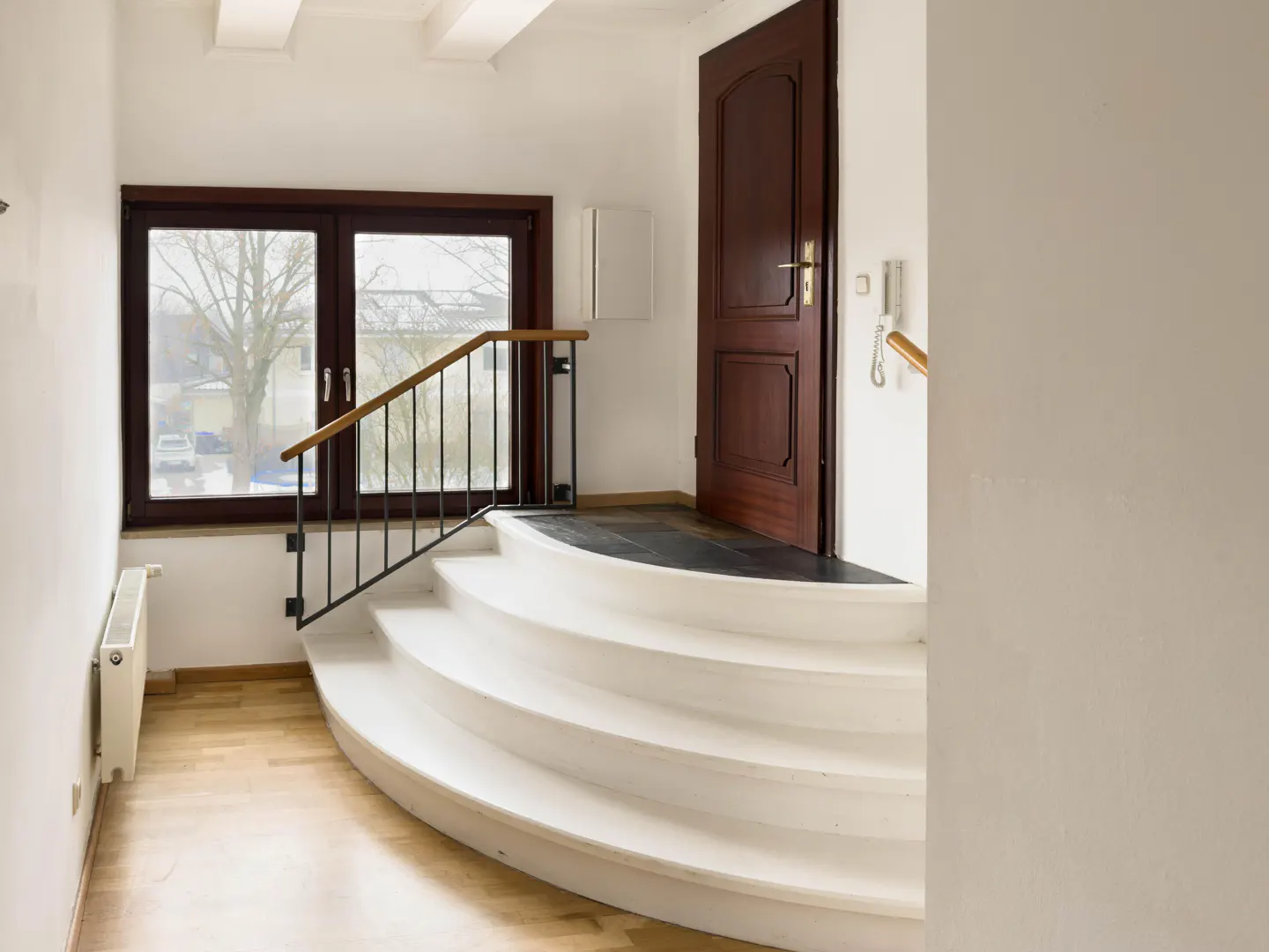 Interior view of a home's entryway with curved white stairs, a dark wood door, and a window with a black railing.