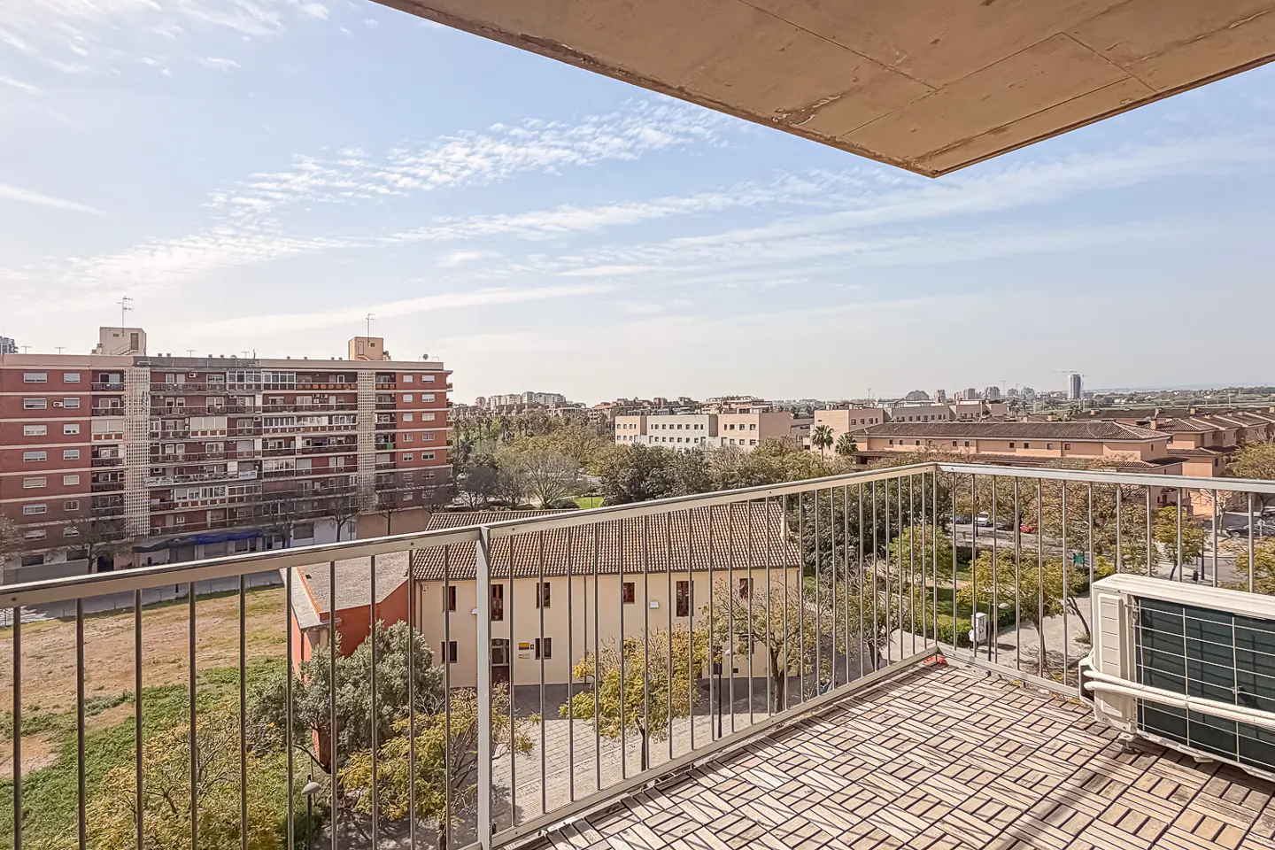 View from a balcony with a metal railing and tiled floor, overlooking buildings and a blue sky with clouds.