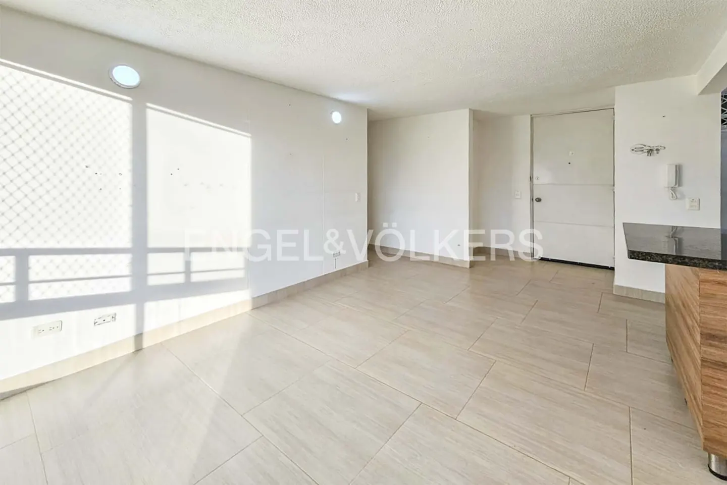 Bright, empty room with white walls, beige tile floor, and a large window with sunlight streaming in. A white door is visible on the right.