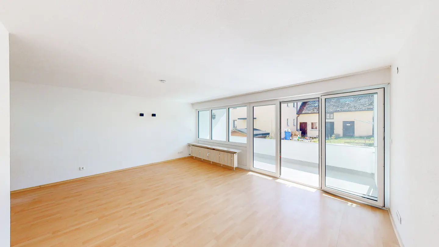 Bright, empty room with light wood floors, white walls, and large sliding glass doors to a balcony. A radiator sits under the windows.