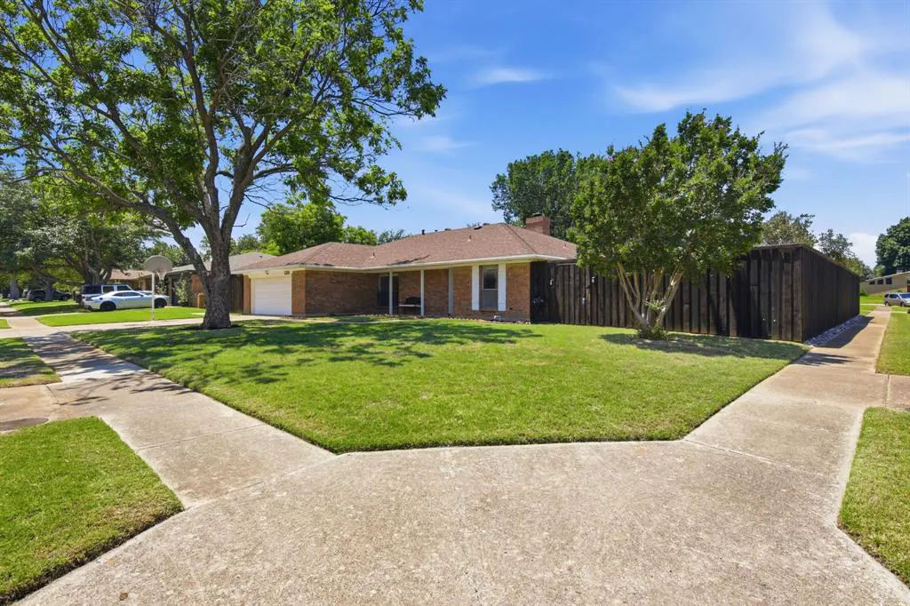A single-story brick house with a brown roof, green lawn, and a large tree in the front yard.