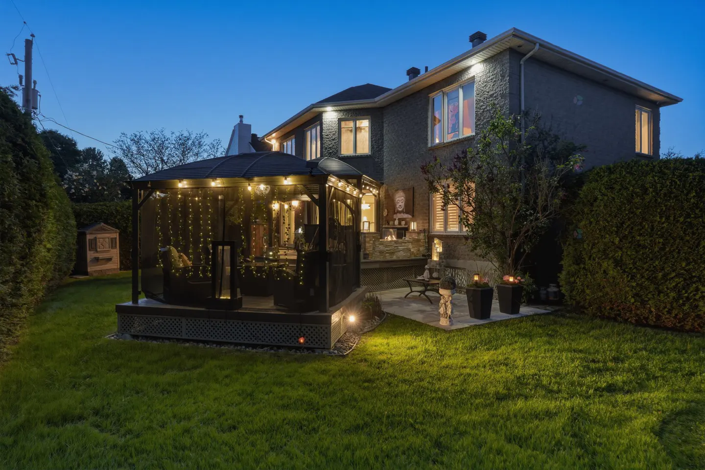 Backyard of a two-story home at dusk, featuring a gazebo with string lights, a stone patio, and a lush green lawn.