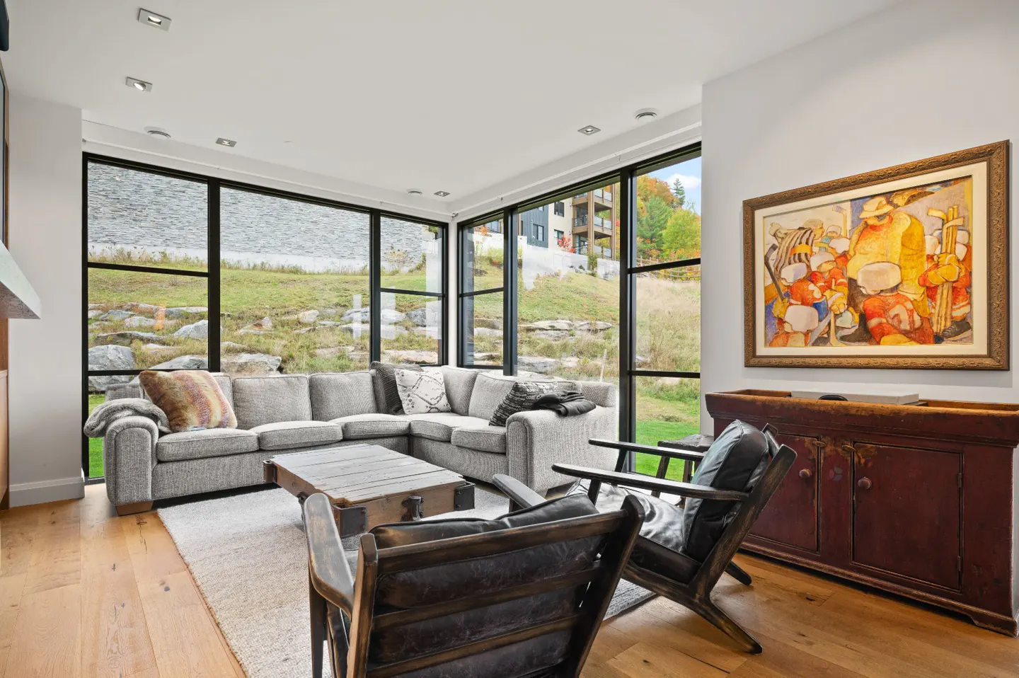 Living room with gray sectional, wood chairs, and large windows overlooking a grassy hill. A painting hangs above a red cabinet.