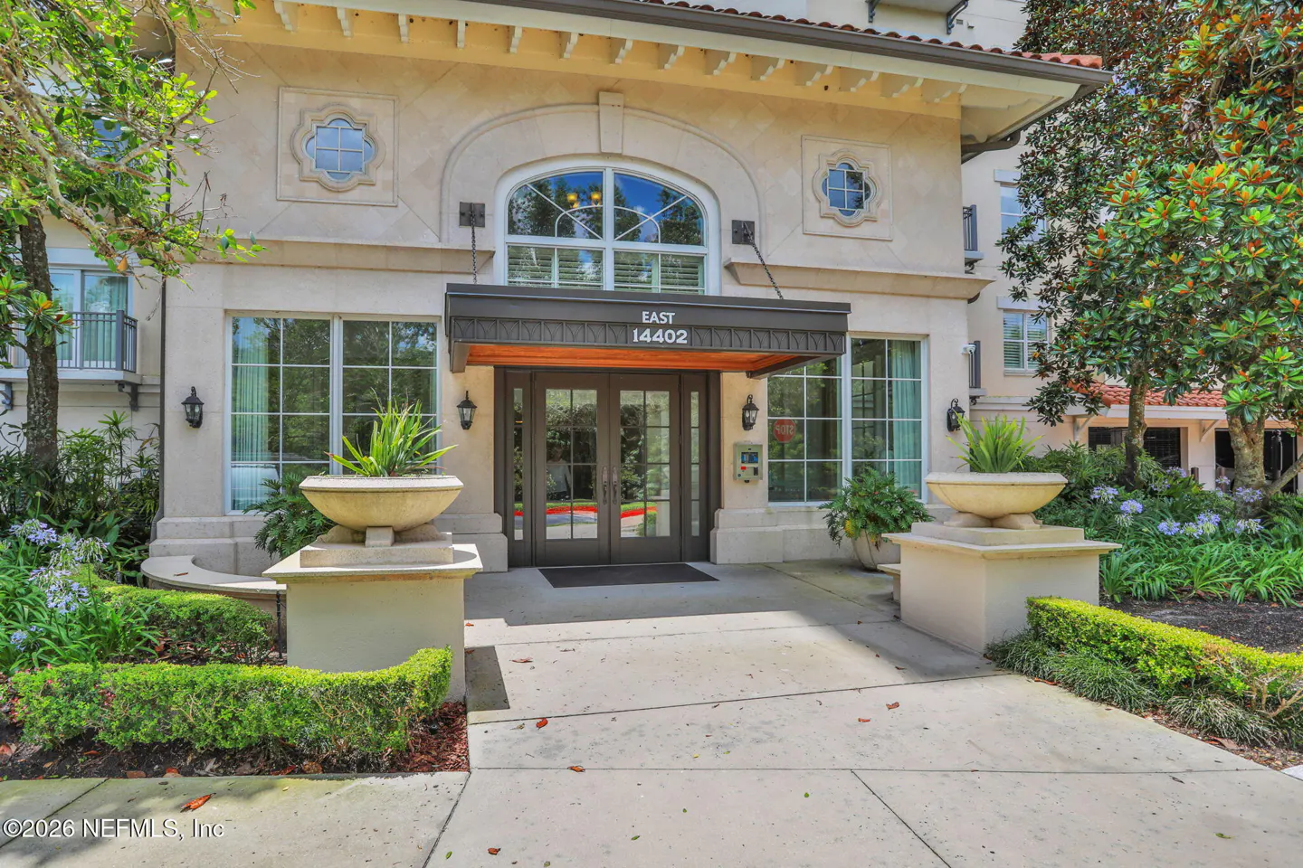 Exterior view of a beige building with a glass door entrance. A black awning reads "EAST 14402". Planters and manicured hedges flank the walkway.