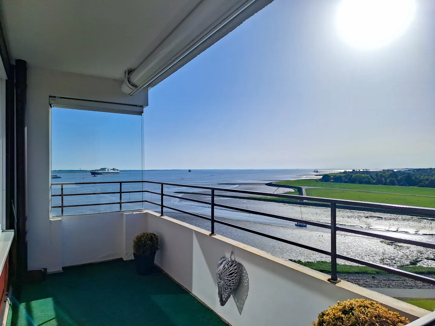 Balcony view overlooking a river with a cargo ship, green fields, and bright sunlight under a clear blue sky.