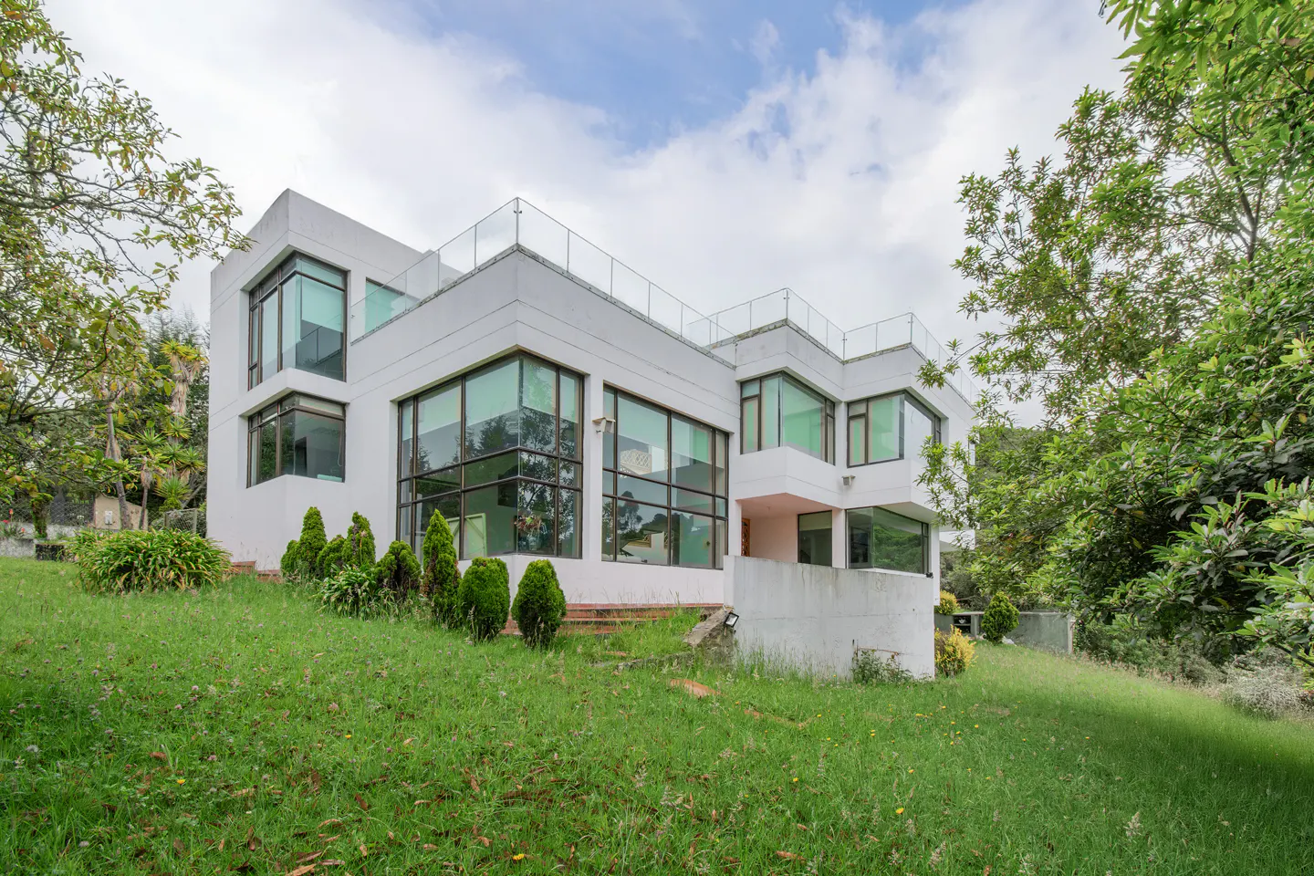 Modern white house with large windows and a glass-railed rooftop terrace, set on a green lawn with trees.