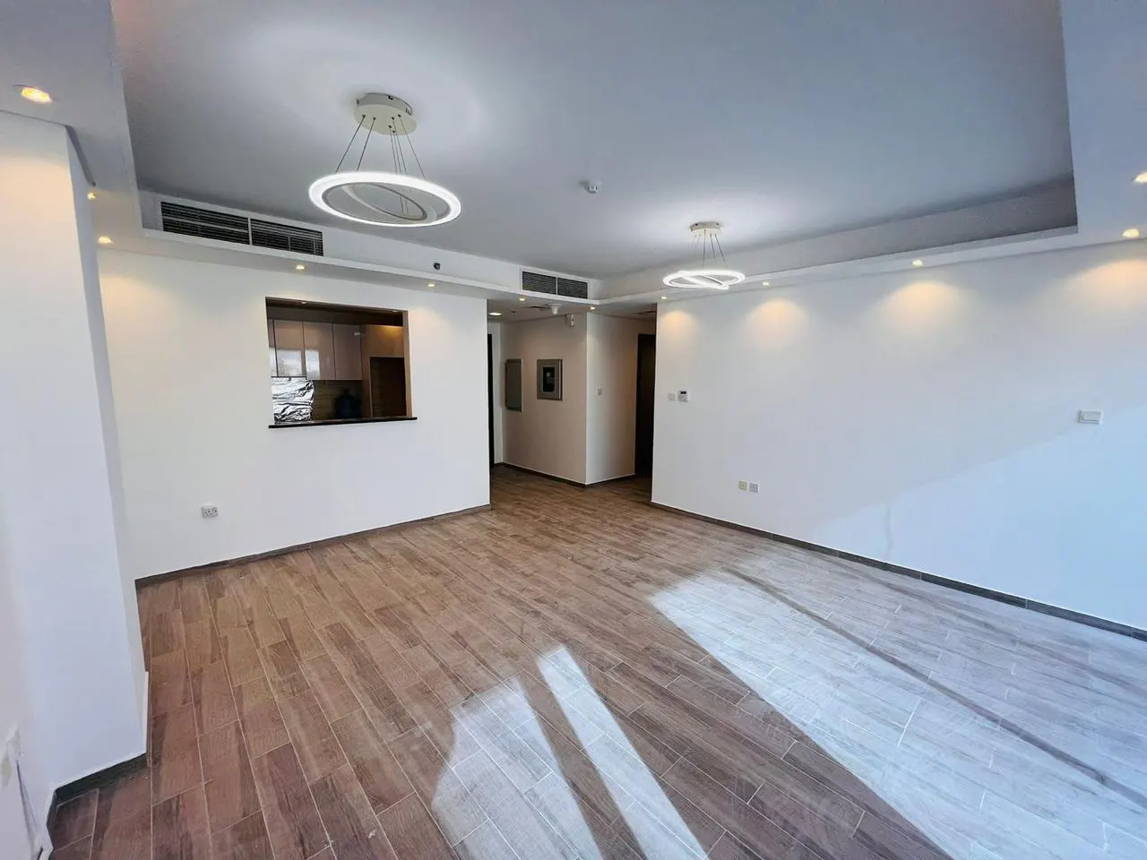Spacious living room with white walls, wood-look tile floors, modern circular pendant lights, and a kitchen pass-through.