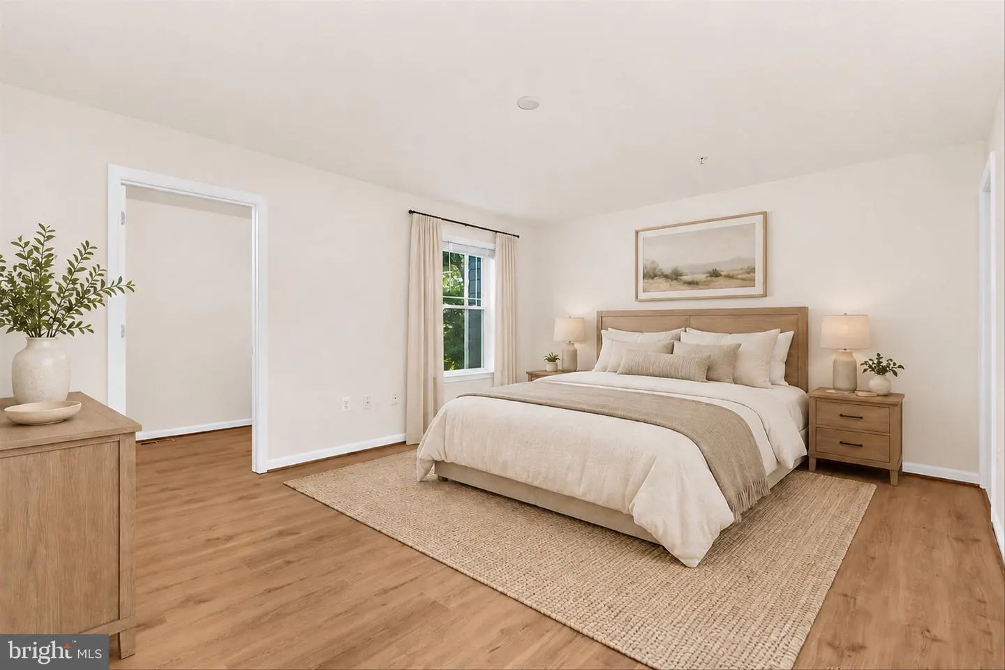 Bedroom with light wood floors, beige walls, and a large bed with neutral bedding. A window with curtains is visible.