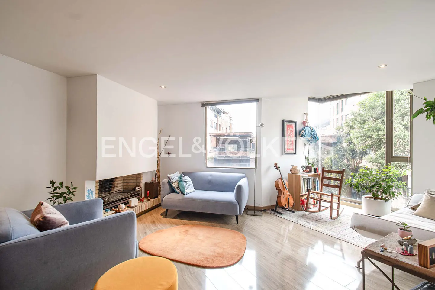 Bright living room with gray sofas, a fireplace, and a large window overlooking a city street. A violin and rocking chair add character.
