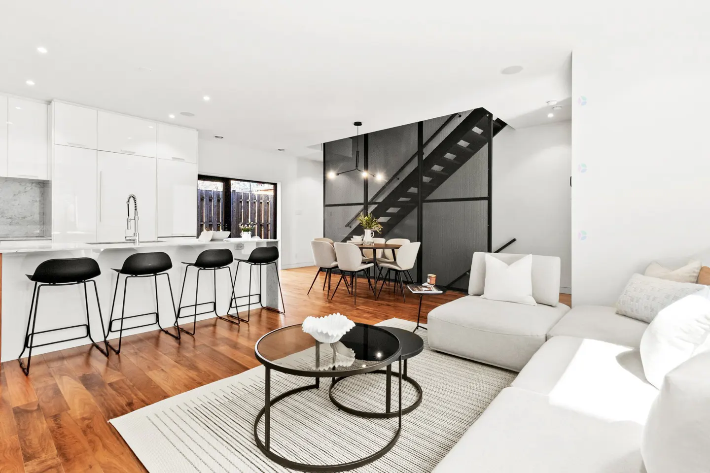 Modern open-concept living area with white kitchen, black bar stools, dining set, and a light gray sectional sofa.