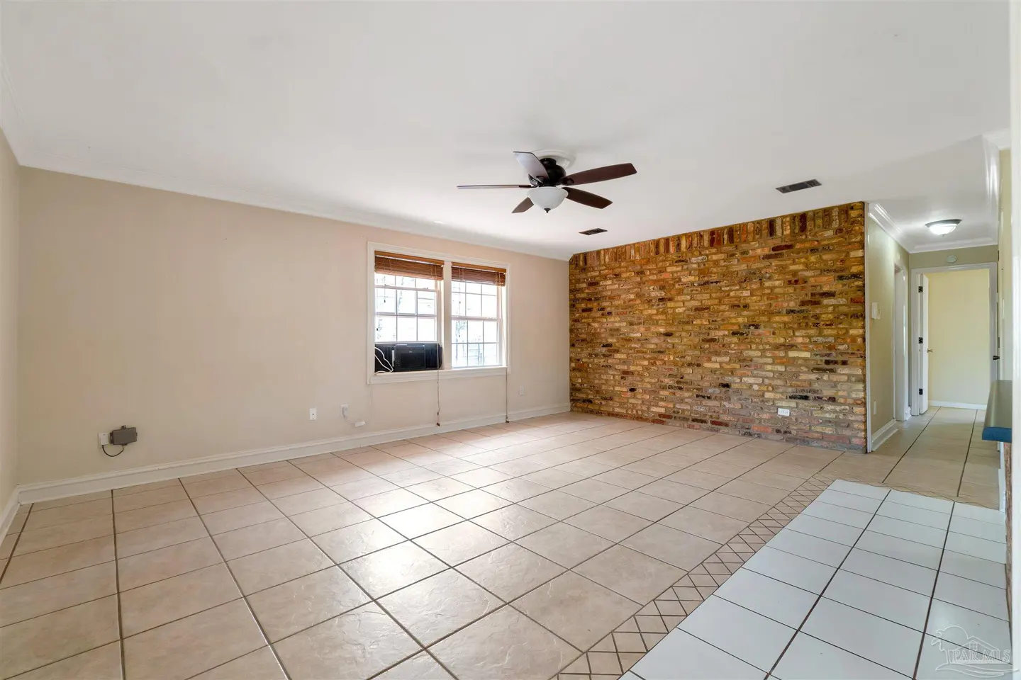 A spacious living room with beige tile flooring, a brick accent wall, and a ceiling fan. Natural light streams through the window.