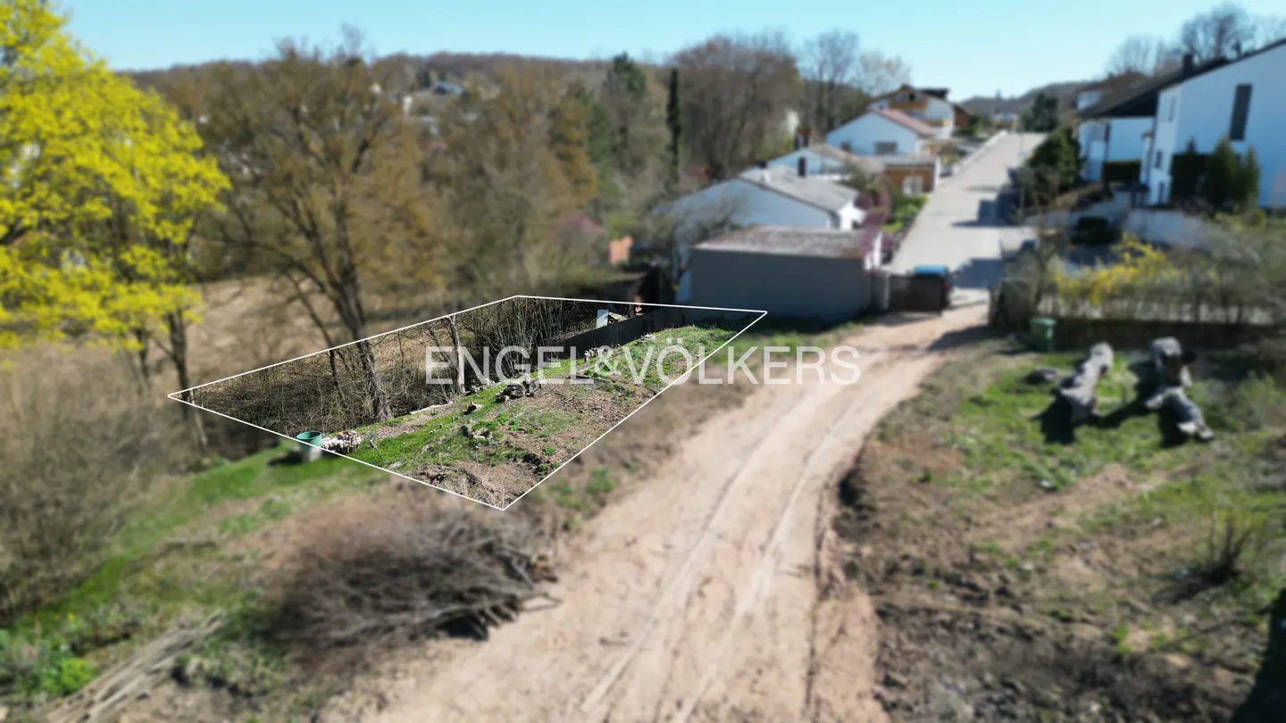 Aerial view of a vacant lot outlined in white, with trees, houses, and a dirt road in the background. "Engel & Völkers" is overlaid.