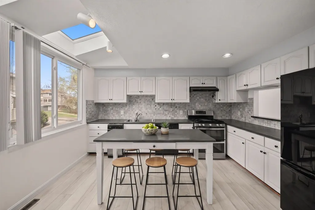 Bright kitchen with white cabinets, black countertops, and a gray island with three wooden stools. A large window and skylight provide natural light.