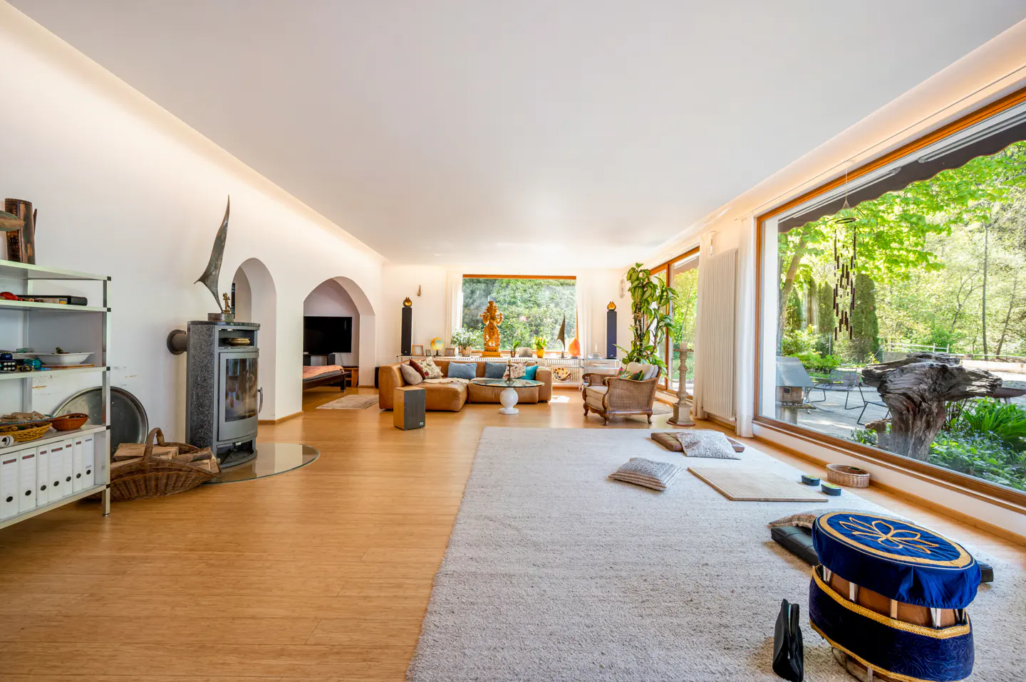 Bright living room with wood floors, white walls, and large windows overlooking a green garden. A brown leather sofa and a fireplace add warmth.