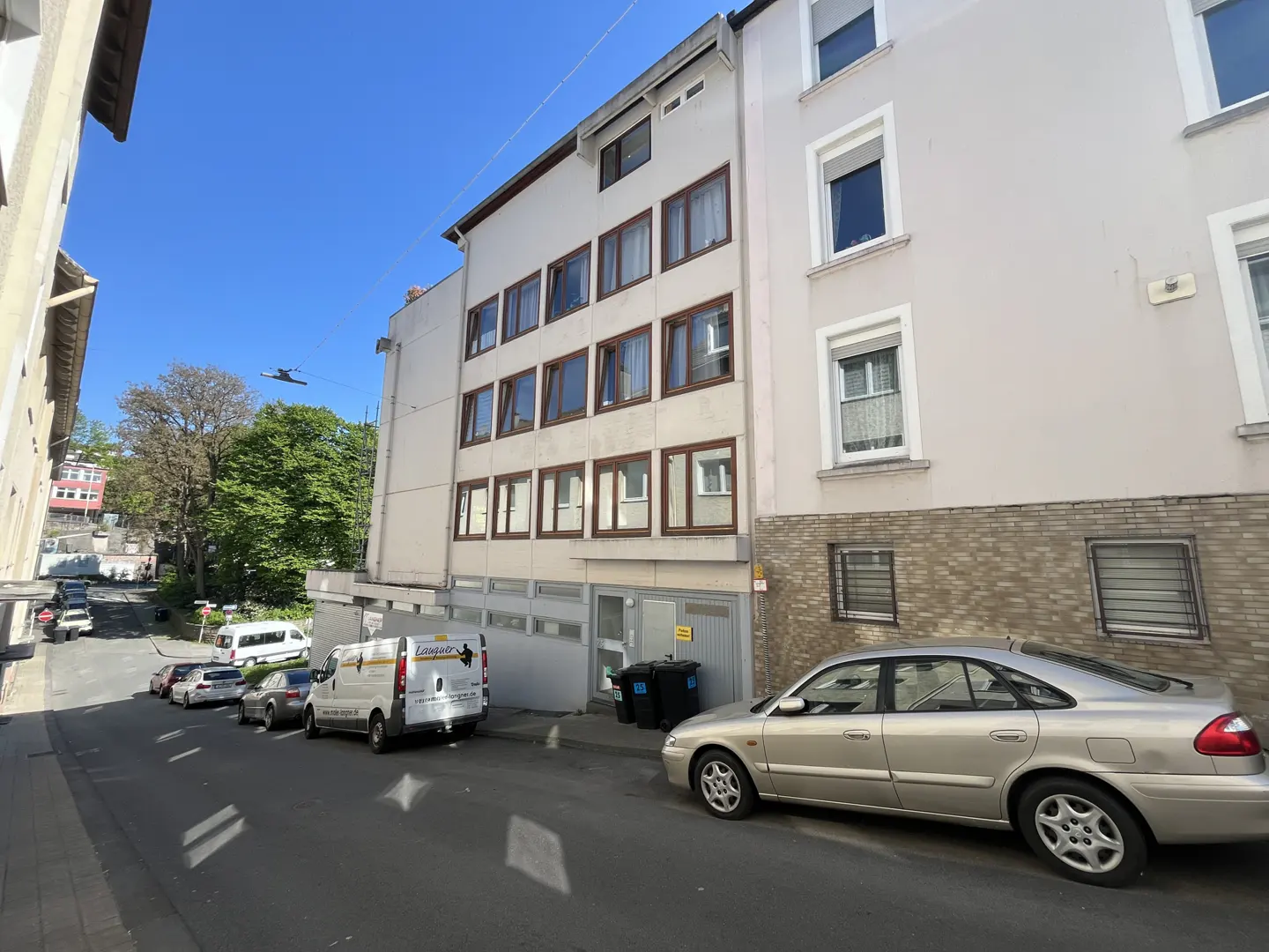 Street view of a multi-story building with brown-framed windows. Cars and a white van are parked along the street. Blue sky is visible.