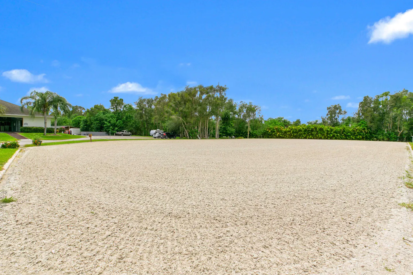 A large, empty sand arena under a blue sky, surrounded by green trees and a building in the background.