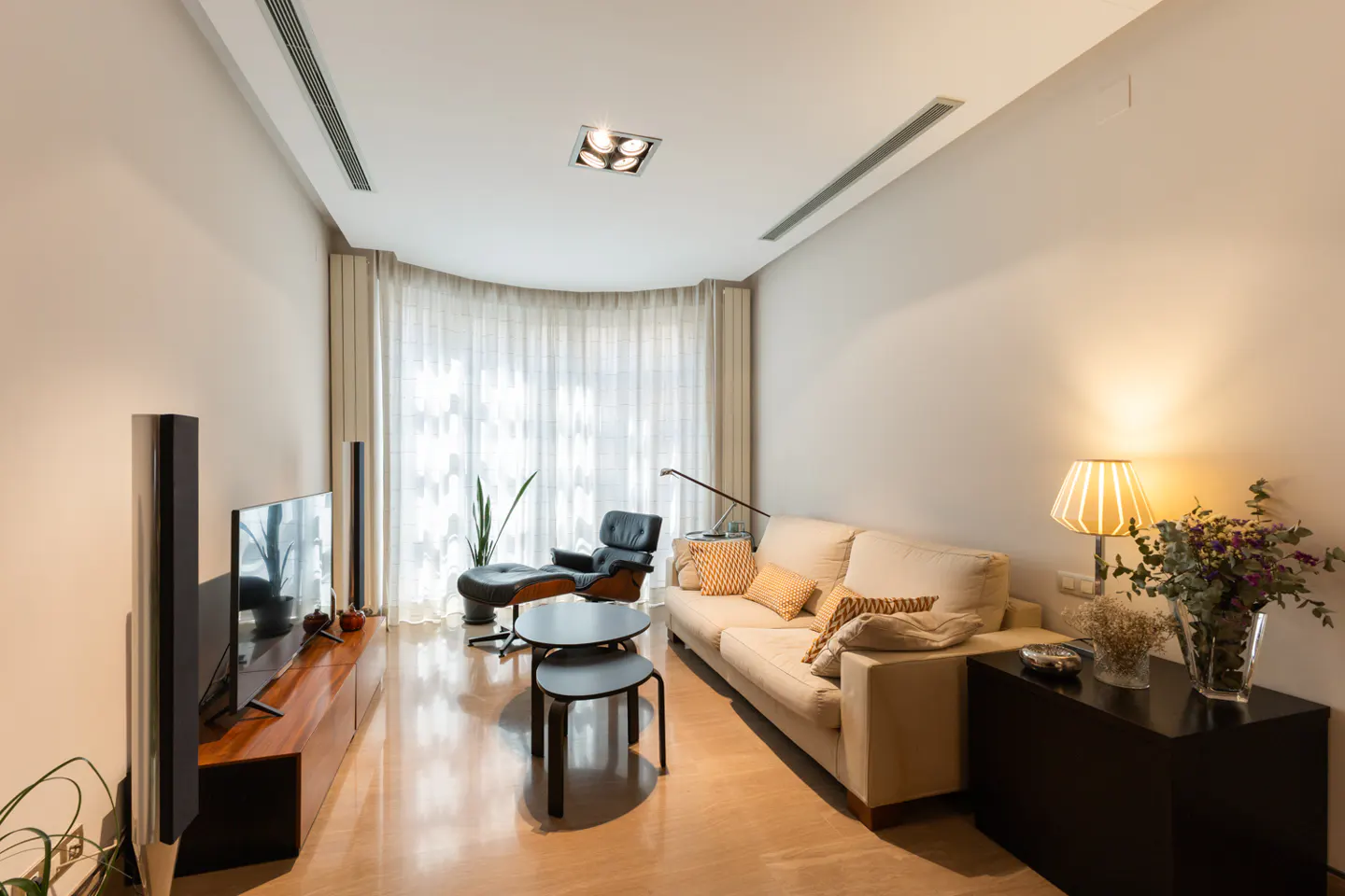 Living room with beige walls, light wood floors, and a cream sofa with patterned pillows. A black leather lounge chair sits near sheer curtains.