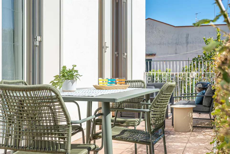 Outdoor patio with green table and chairs, colorful glasses, and a potted plant. Lounge seating is visible in the background.