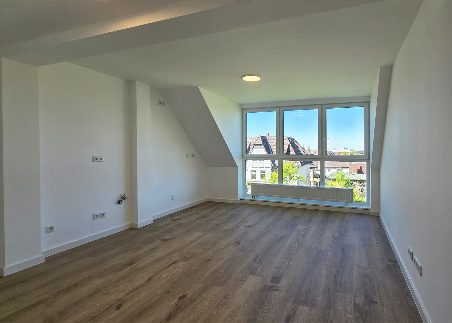 Bright, empty room with wood floors, white walls, and a large window showing a neighborhood view.