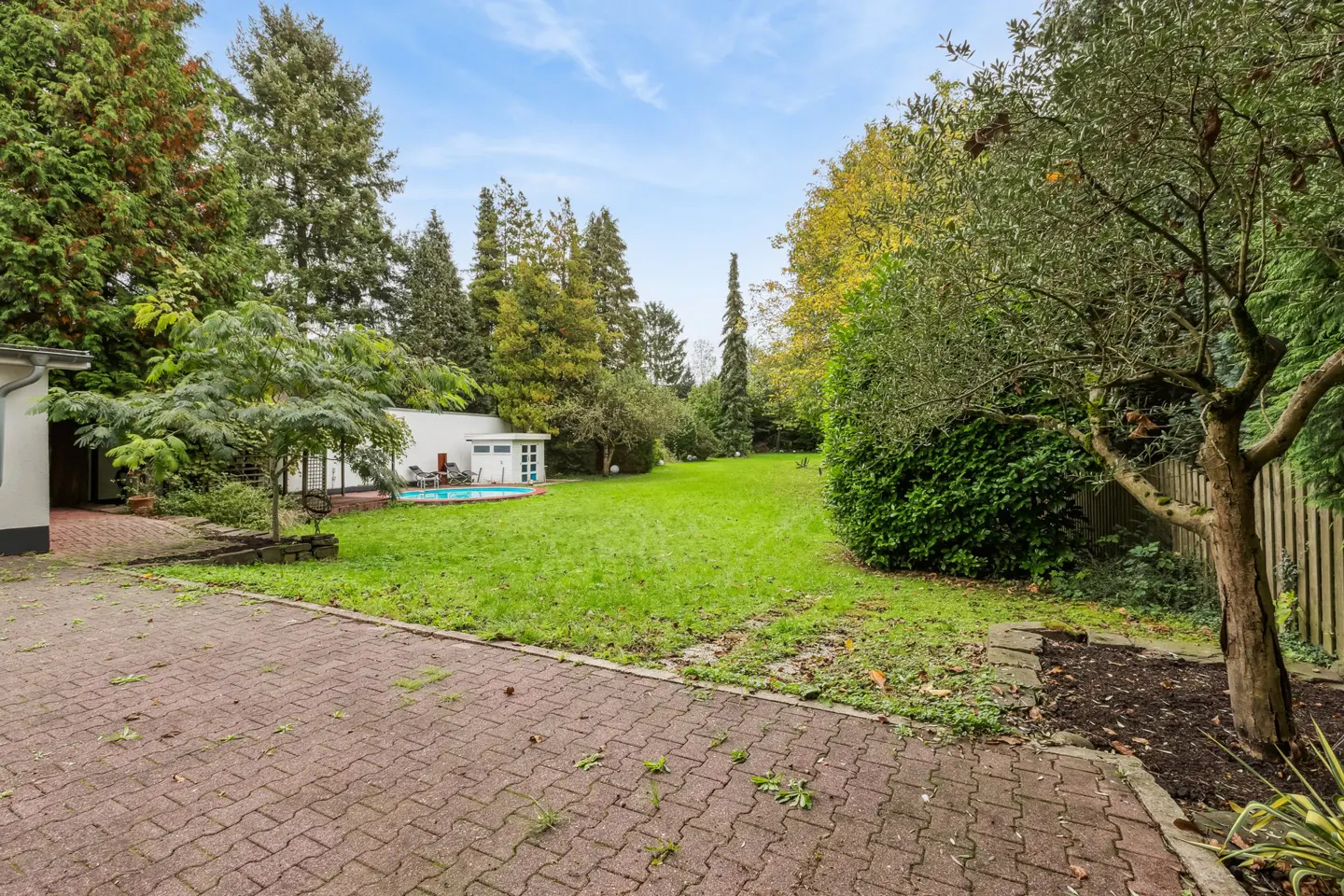 A backyard with a brick patio, green lawn, trees, and a pool with a white pool house.