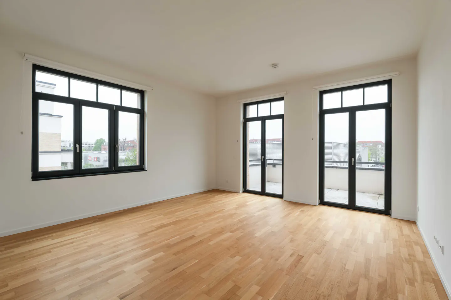 Bright, empty room with light wood floors, white walls, and black-framed windows and doors leading to balconies.
