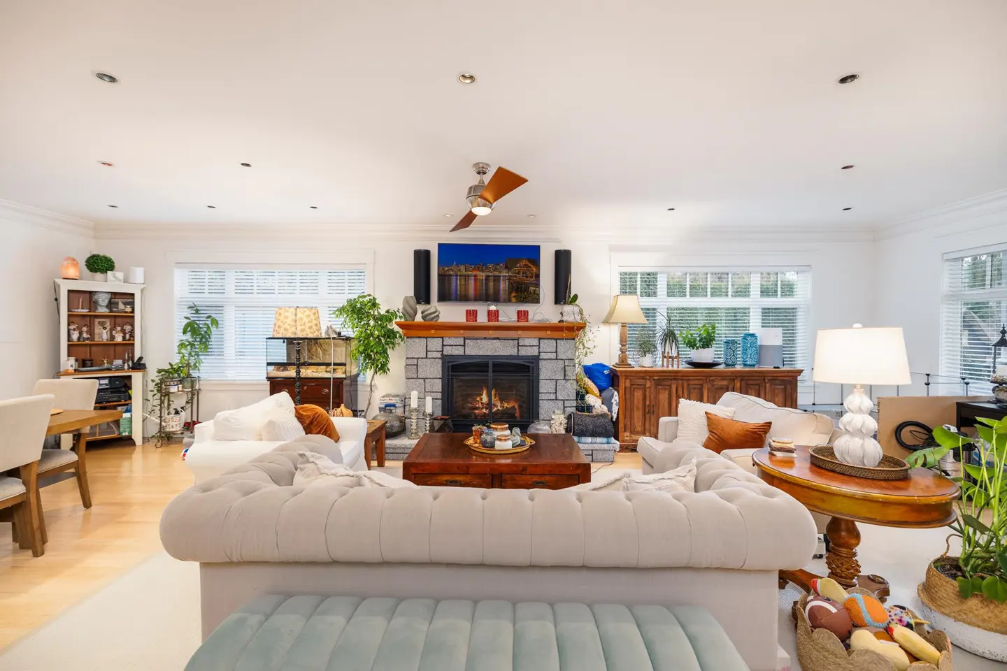 Bright living room with a stone fireplace, white sofas, wood floors, and a ceiling fan. A TV hangs above the mantel.