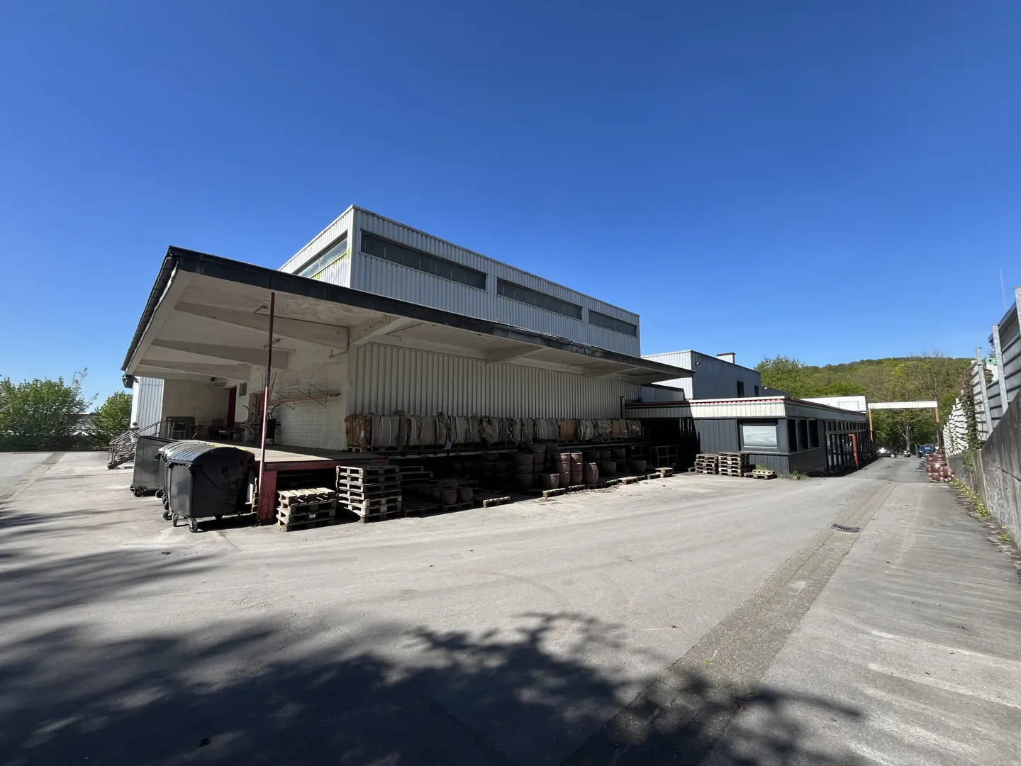 Exterior view of a white industrial building with a loading dock, pallets, and barrels under a blue sky.