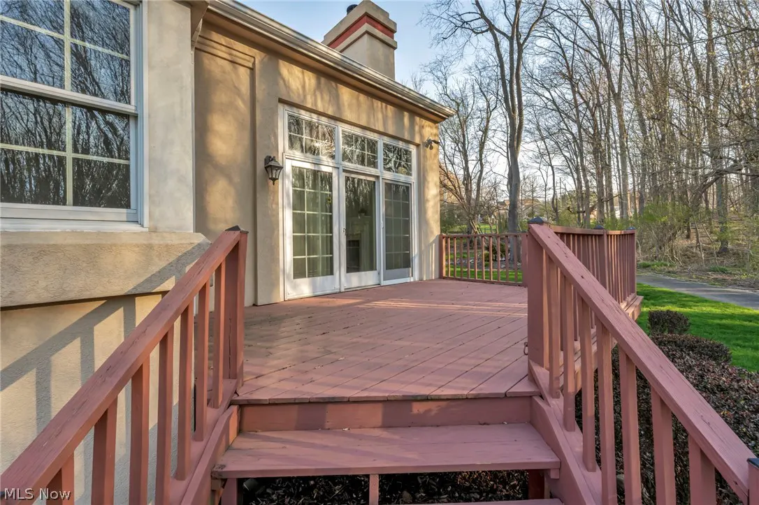 Exterior view of a tan house with a red wood deck, stairs, and white framed glass doors. Trees are visible in the background.