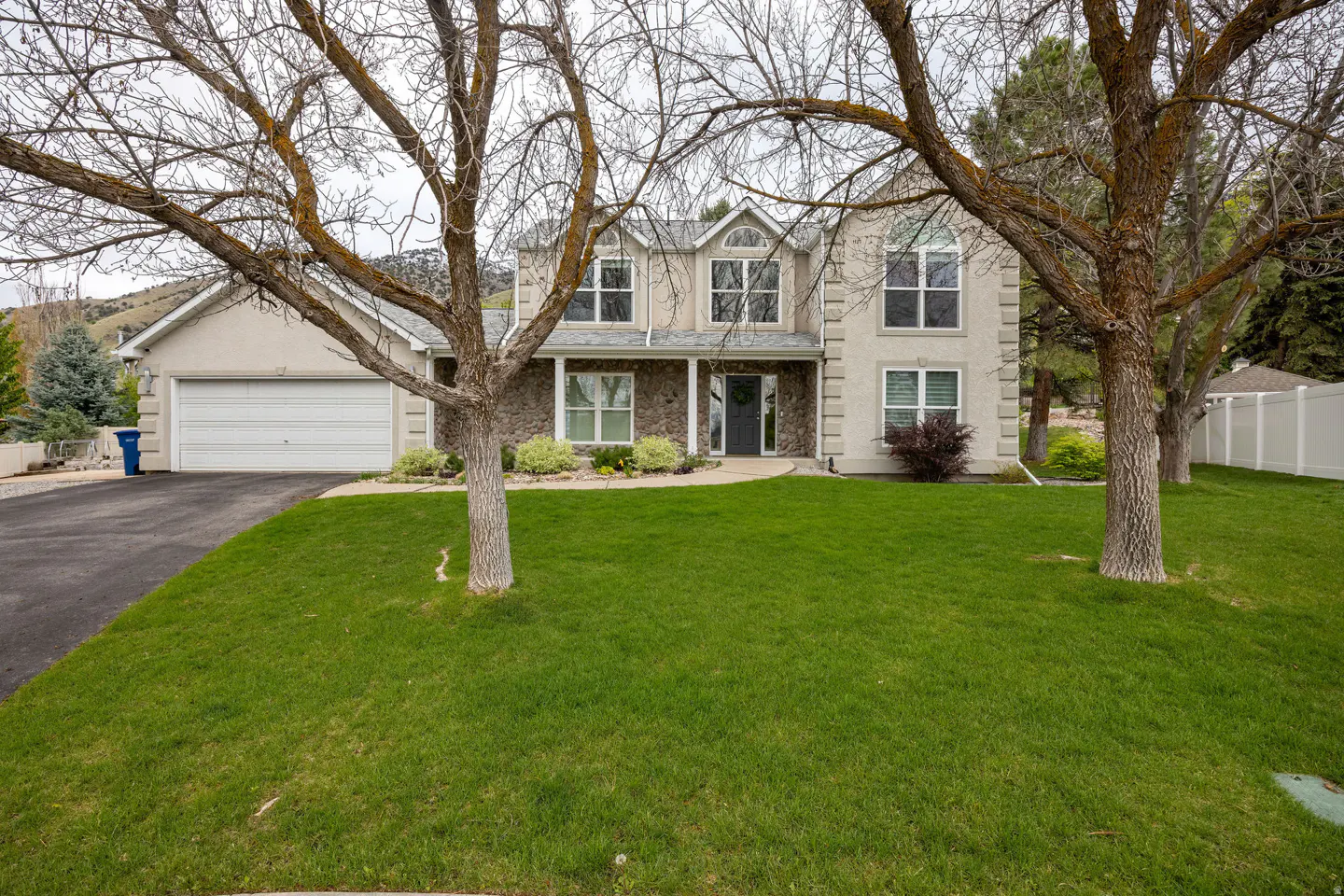 Two-story house with stone and stucco facade, large lawn, and trees in the foreground. Garage on the left, mountain in the background.