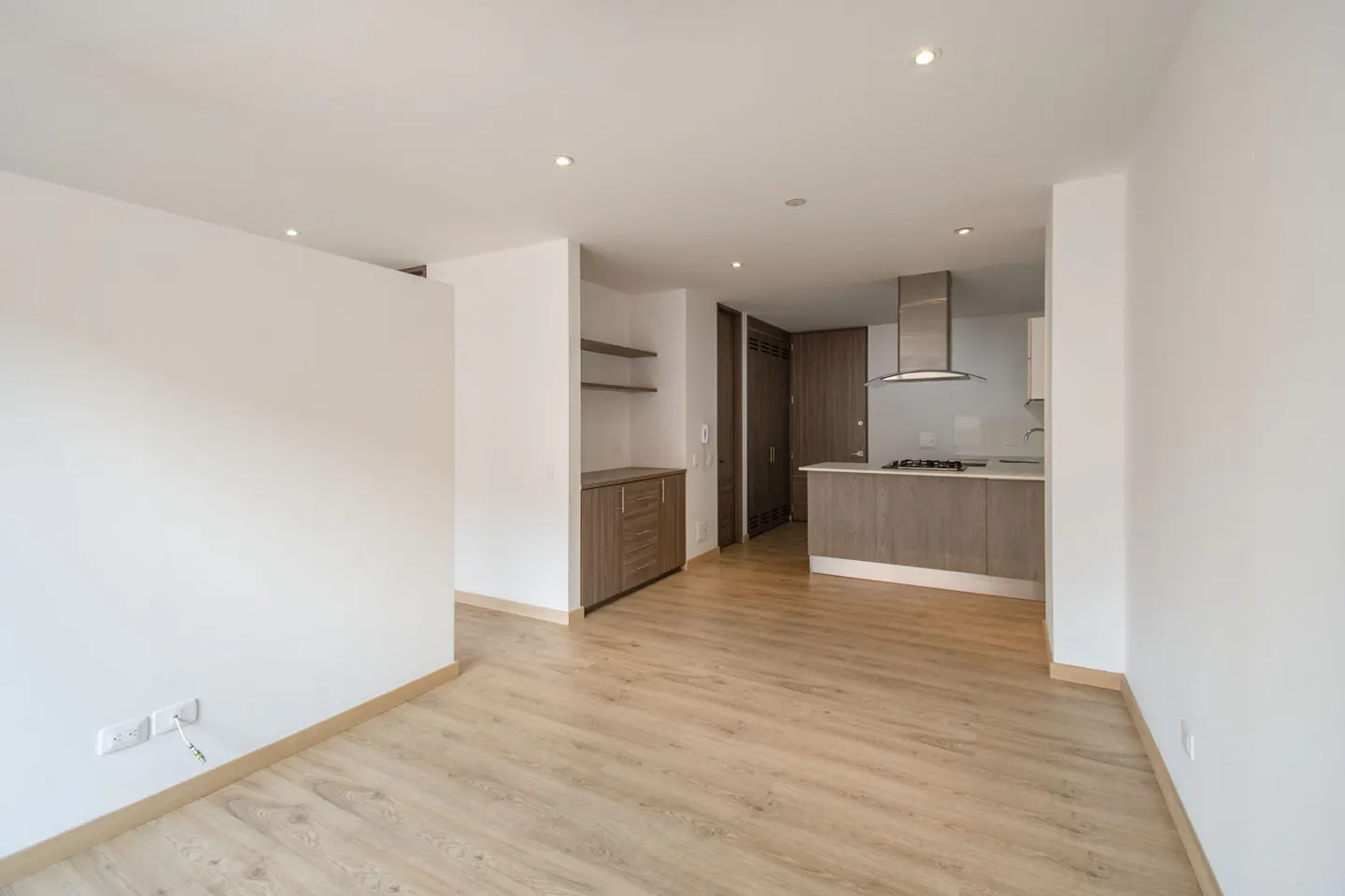 Bright, empty apartment with light wood floors, white walls, and recessed lighting. A kitchen with stainless steel hood and wood cabinets is visible.