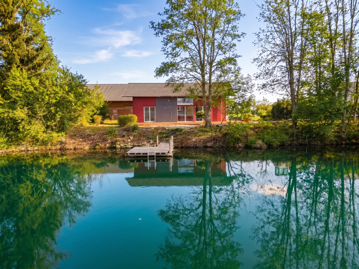 Modern red and gray house with a dock on a turquoise lake, reflecting trees and the house.