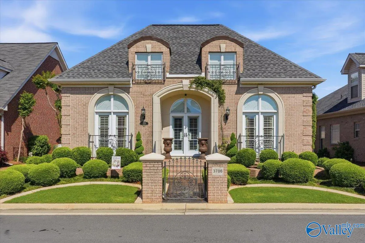 A tan brick house with a gray roof, white trim, and a black iron gate. Green bushes line the front of the house.