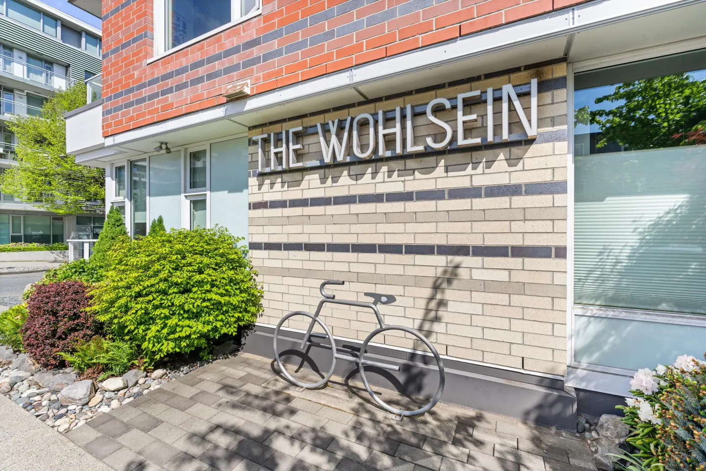Exterior of "The Wohlsein" building with brick facade, bike rack, and landscaping.