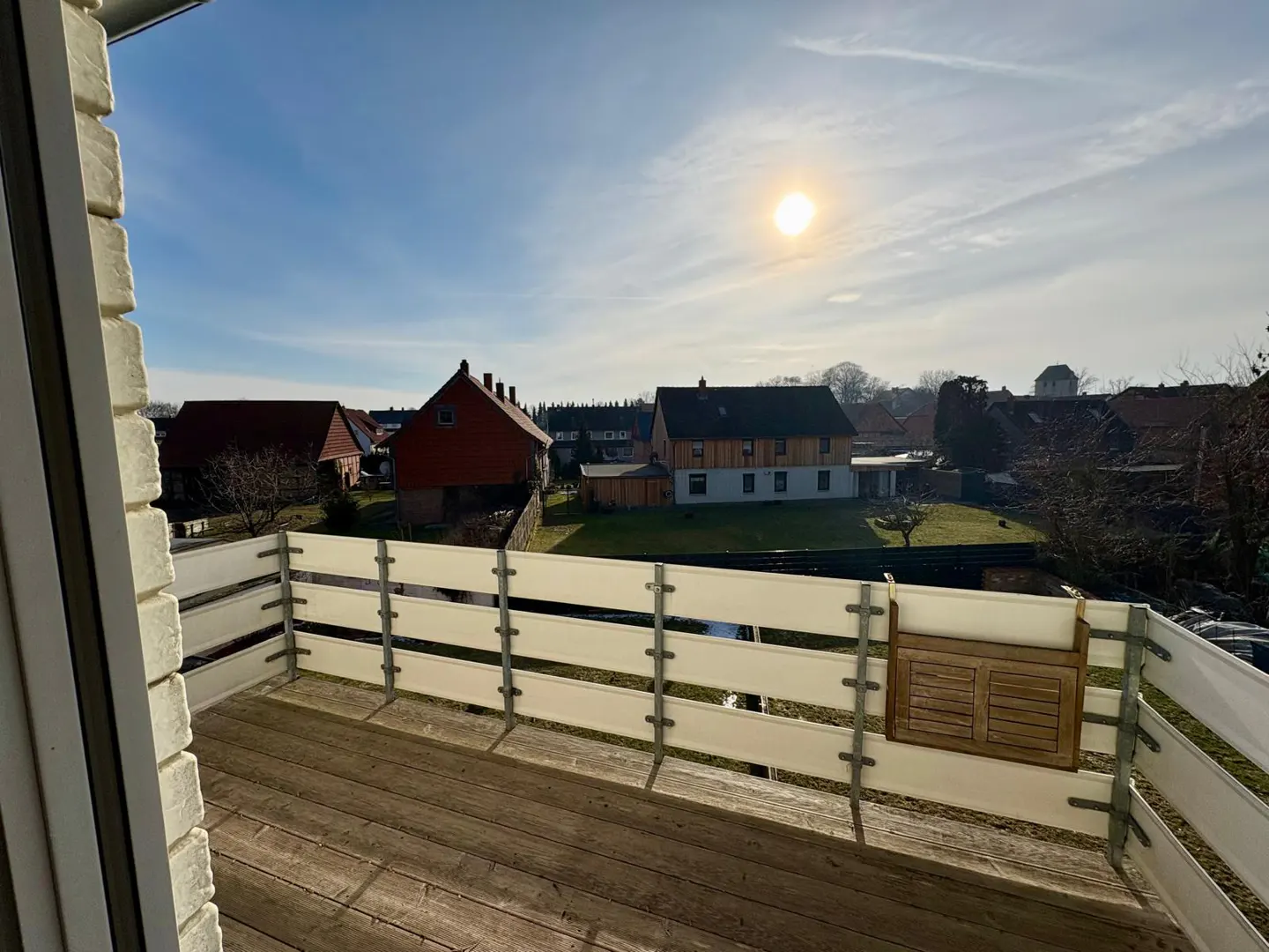 View from a wooden balcony with a white railing, overlooking houses under a blue sky with a bright sun.