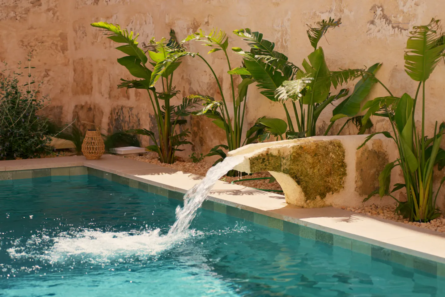 A turquoise pool with a stone fountain and tropical plants against a stone wall.