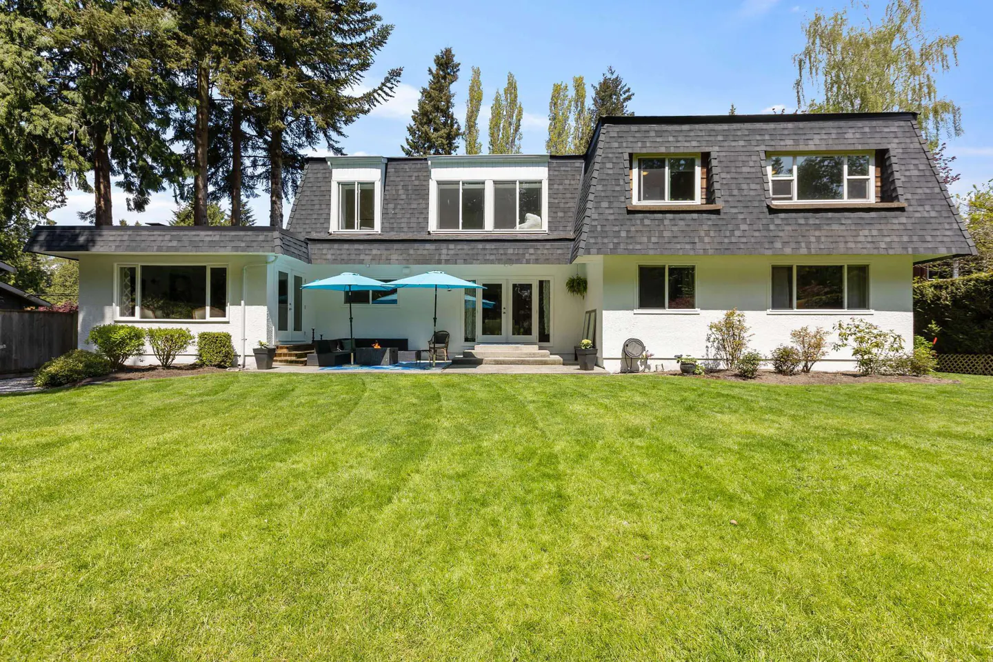 Back of a white house with a dark roof, a green lawn, and a patio with blue umbrellas.
