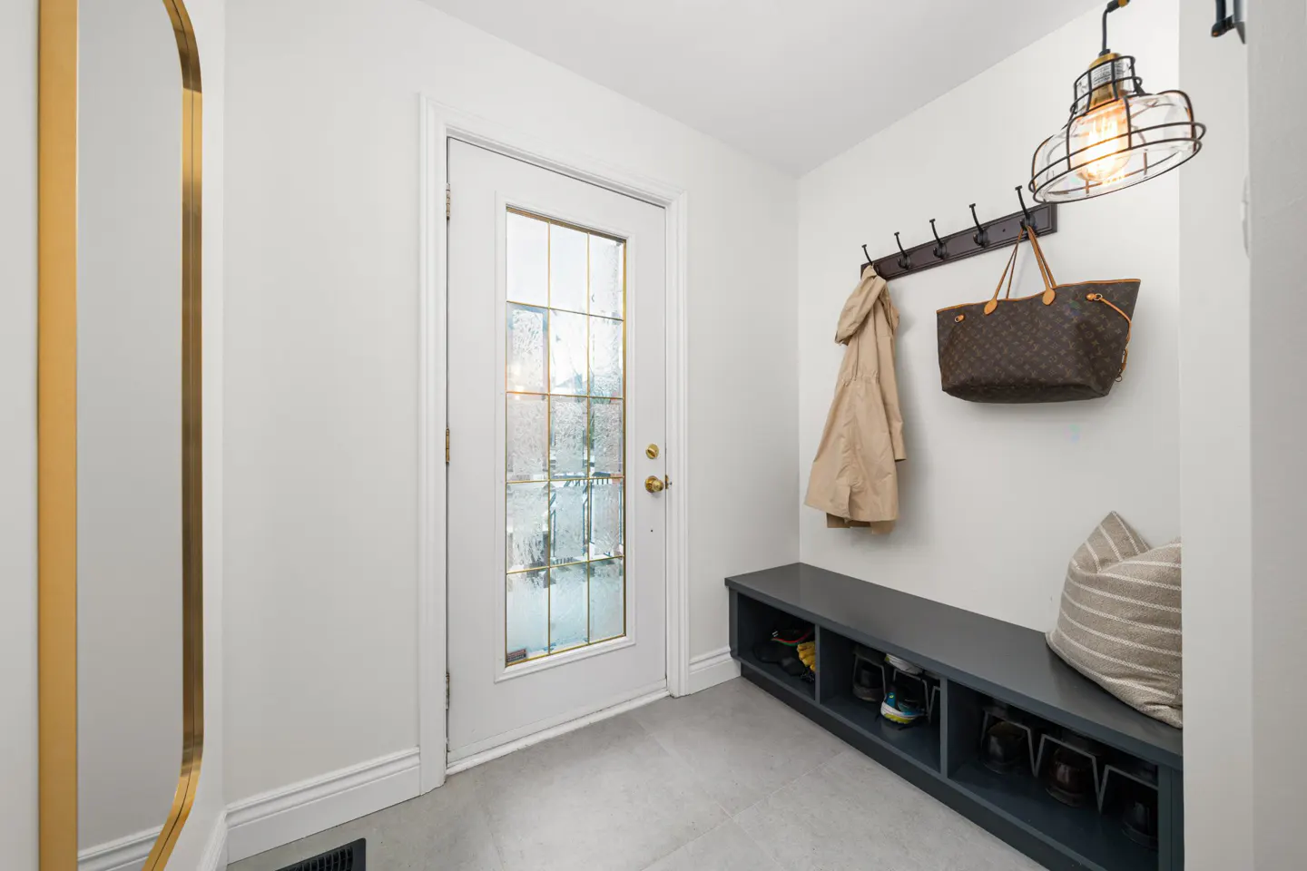 Entryway with a white door, coat rack with a tan coat and purse, a gray bench with shoes, and a gold-framed mirror.