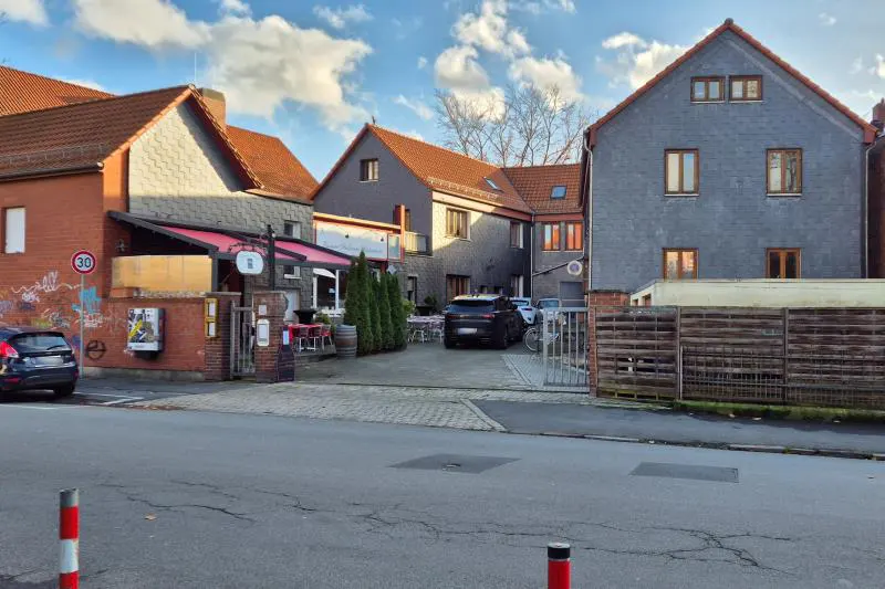 Street view of a restaurant with outdoor seating, surrounded by brick and gray buildings under a partly cloudy sky.