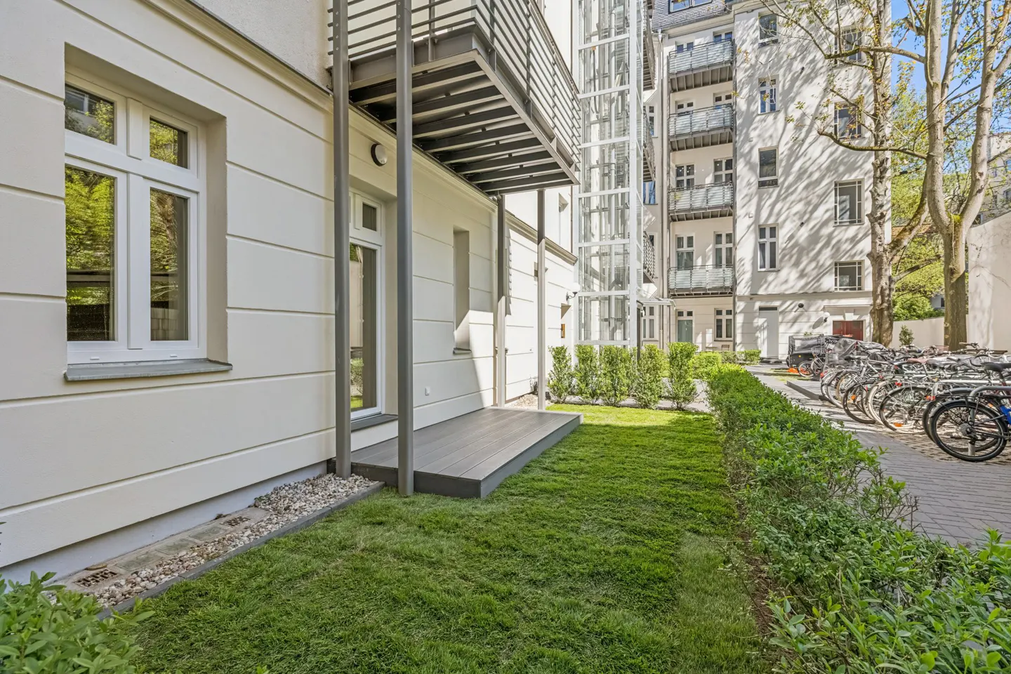 Exterior view of a white building with a small gray deck, green lawn, and a bicycle parking area in the background.