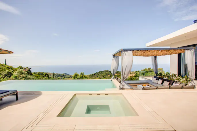 Infinity pool and sunken hot tub on a patio with a shaded lounge area overlooking a scenic ocean and hillside view.