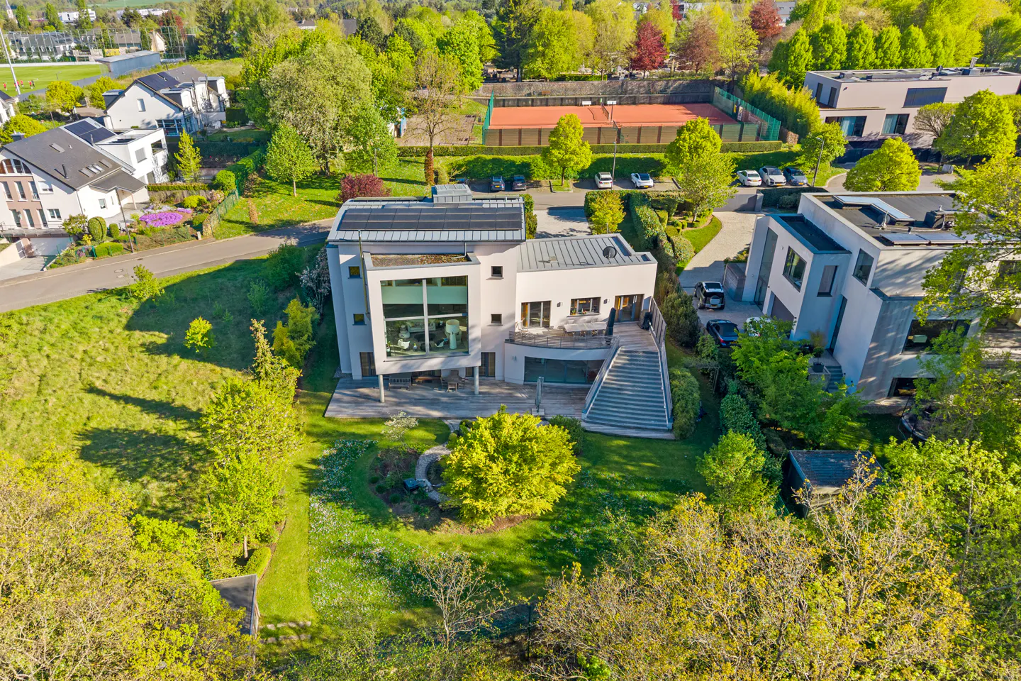 Aerial view of a modern, light gray house with solar panels, a large window, and a staircase leading to a patio, surrounded by green trees.