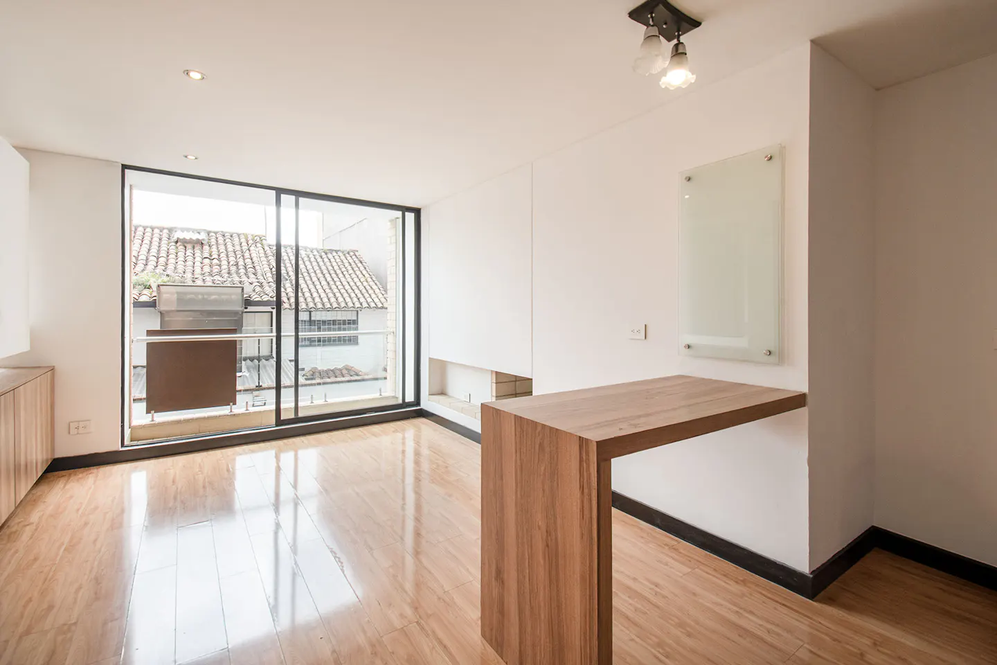 Bright, empty room with wood floors, white walls, and a large window. A wood table is attached to the wall. A glass board hangs above it.