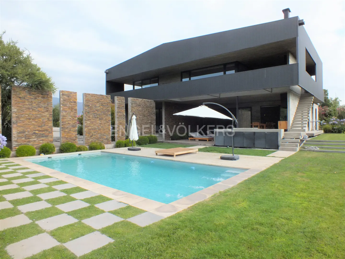 Modern two-story house with a pool. The house is black and gray with a stone wall. The pool is turquoise and surrounded by grass.
