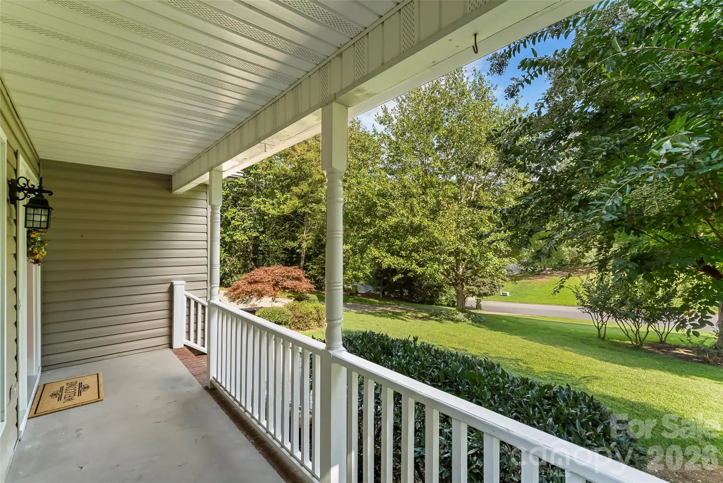 Covered porch with white railings overlooks a green lawn and trees. A welcome mat sits by the front door.