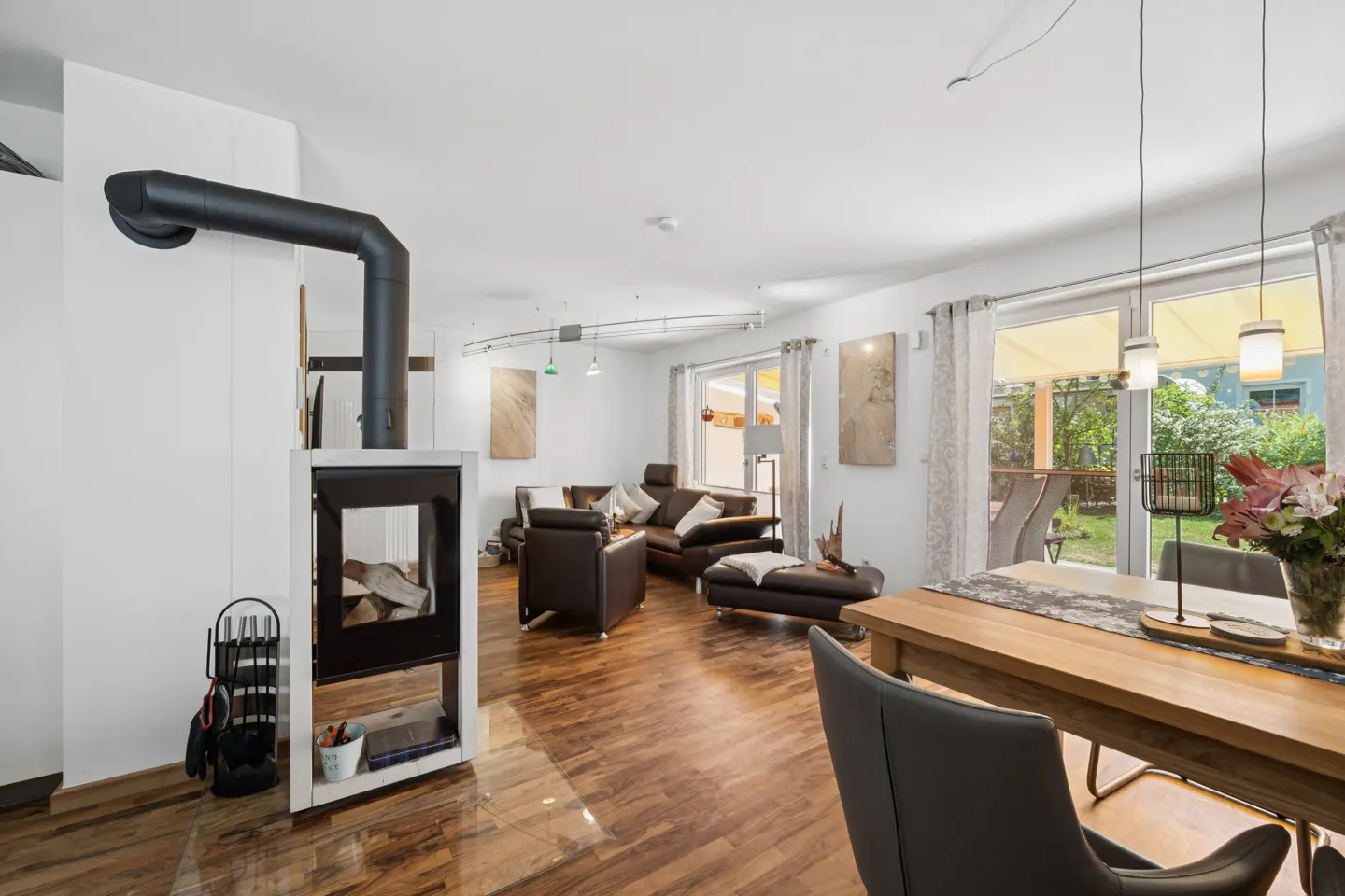 Bright living room with wood floors, a modern fireplace, and brown leather furniture. A dining table sits near a window overlooking a green yard.