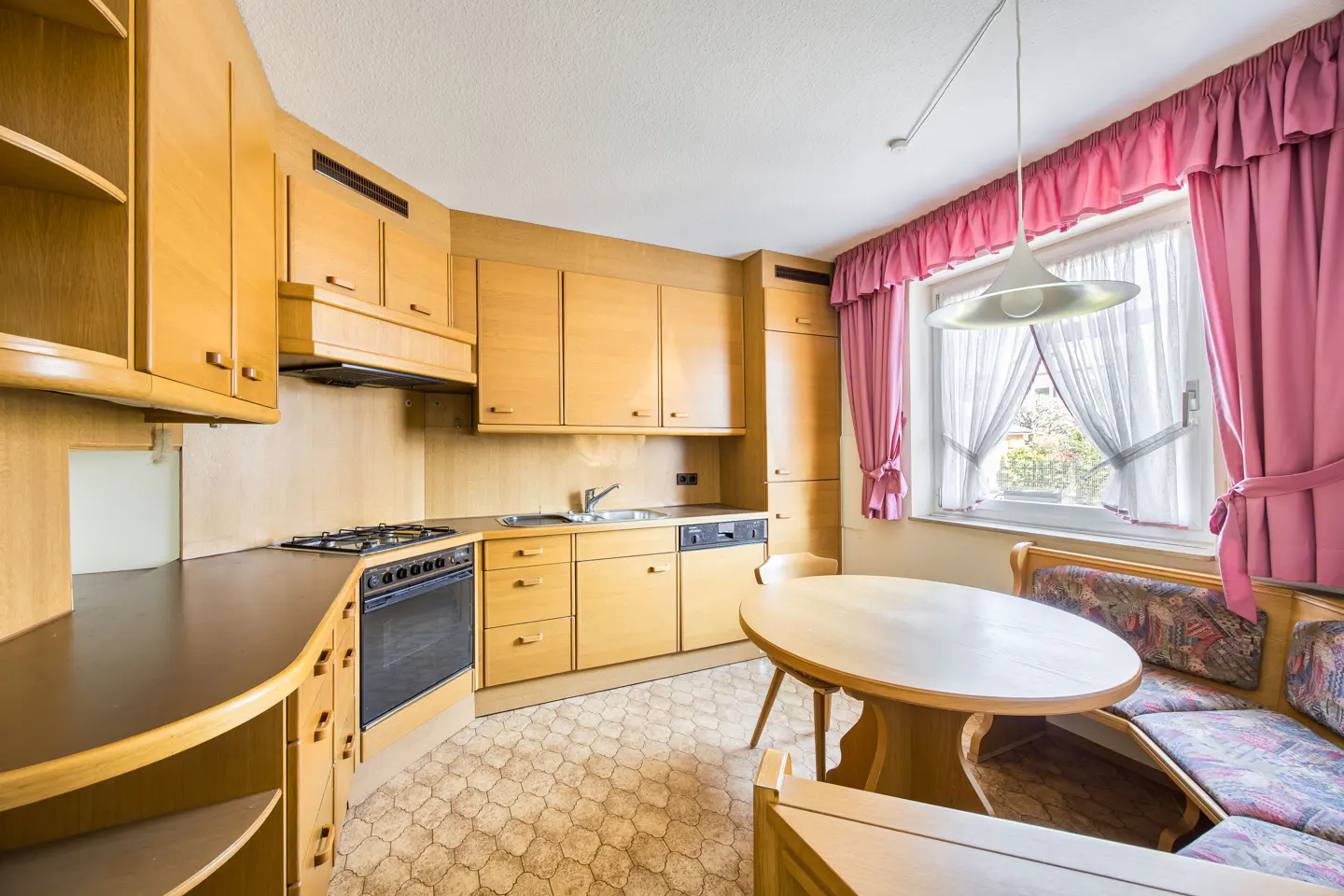 A kitchen with light wood cabinets, a gas stove, and a round table with a cushioned bench. Pink curtains frame a window.